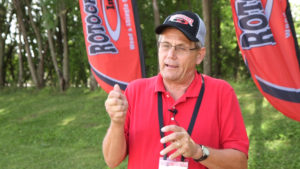 A man outdoors, wearing a hat and speaking with two flags behind him that each say "Rotochopper."