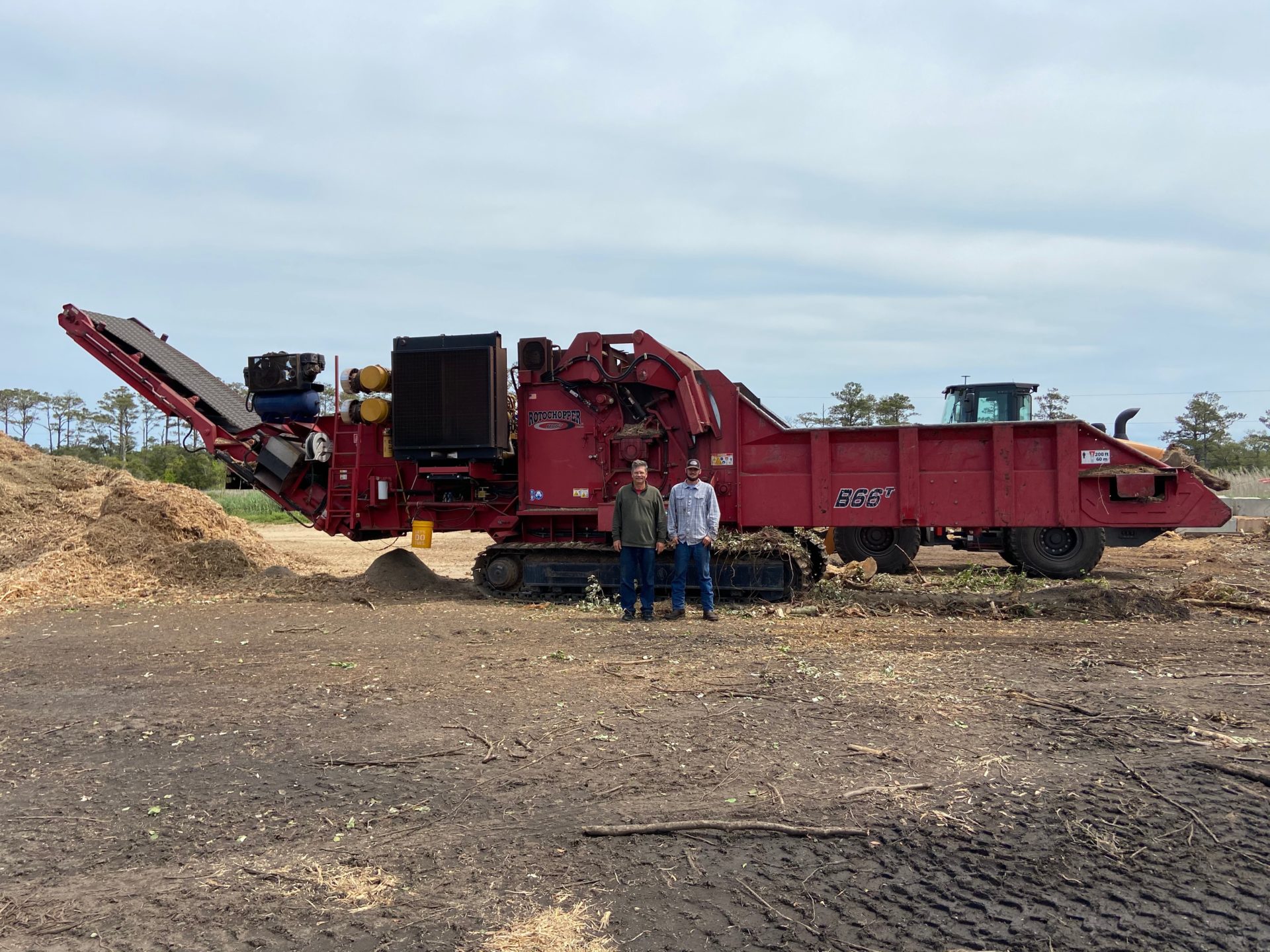 Two men standing in front of a large, red, tracked Rotochopper B-66T mulch-grinding machine