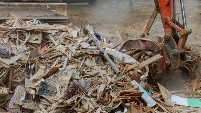 An excavator is shown picking up materials from a large pile of wood and other unsorted C & D materials.