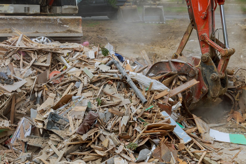 An excavator is shown picking up materials from a large pile of wood and other unsorted C & D materials.