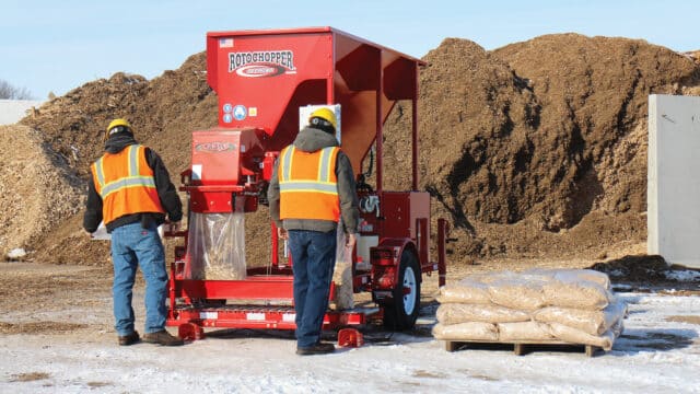 Two operators in high-visibility vests fill and seal bags of mulch using a Rotochopper Go-Bagger 250 at an outdoor yard in winter
