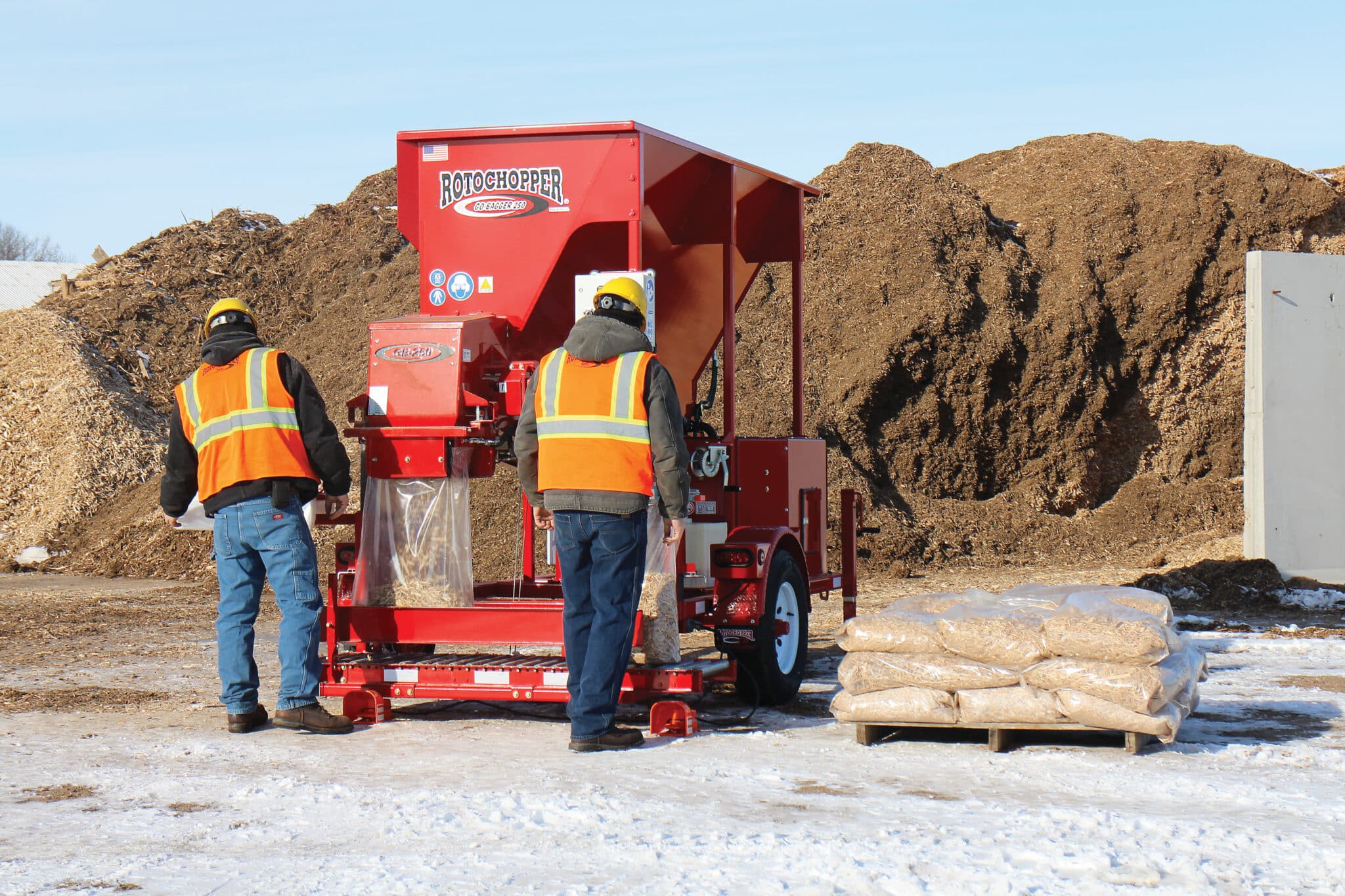 Two operators in high-visibility vests fill and seal bags of mulch using a Rotochopper Go-Bagger 250 at an outdoor yard in winter